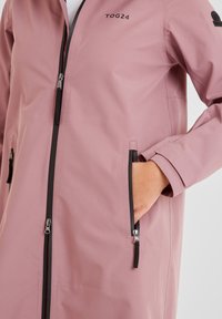 Pink waterproof jacket with a front zipper, black accents, and two side pockets. Features a smooth texture and a relaxed fit design.