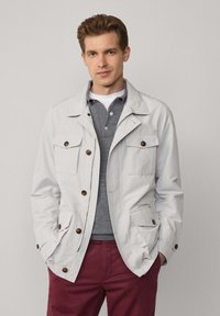 Veste légère gris clair avec quatre poches frontales, accents en bouton marron et col. Portée par-dessus un polo gris.