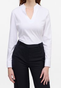 Top bianco a maniche lunghe con scollatura a V poco profonda, abbinato a pantaloni scuri su misura. Il tessuto è liscio e i polsini presentano un classico dettaglio con bottone.