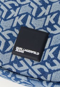 Denim audums ar zilu un gaiši zilu haizivju rakstu, papildināts ar melnu taisnstūra etiķeti, uz kuras baltā krāsā ir uzrakstīts "Karl Lagerfeld Jeans".
