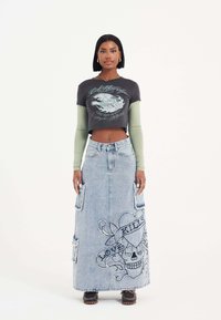 Schwarzes grafisches Crop-Top mit kurzen Ärmeln, das über einem grünen Langarmshirt getragen wird. Helles, blaues Jeans-Maxikleid mit Stickereien von Schädeln und Herzen.