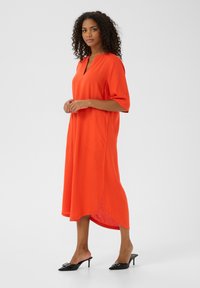 Robe rouge au-dessus du genou avec des manches courtes, un col en V, des poches latérales et un ourlet asymétrique. Porte avec des chaussures à talons noires à enfiler.