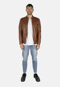 Giacca in pelle marrone con colletto alto, zip frontale, due tasche sul petto, indossata sopra una maglietta bianca, abbinata a jeans azzurri chiari e scarpe nere.