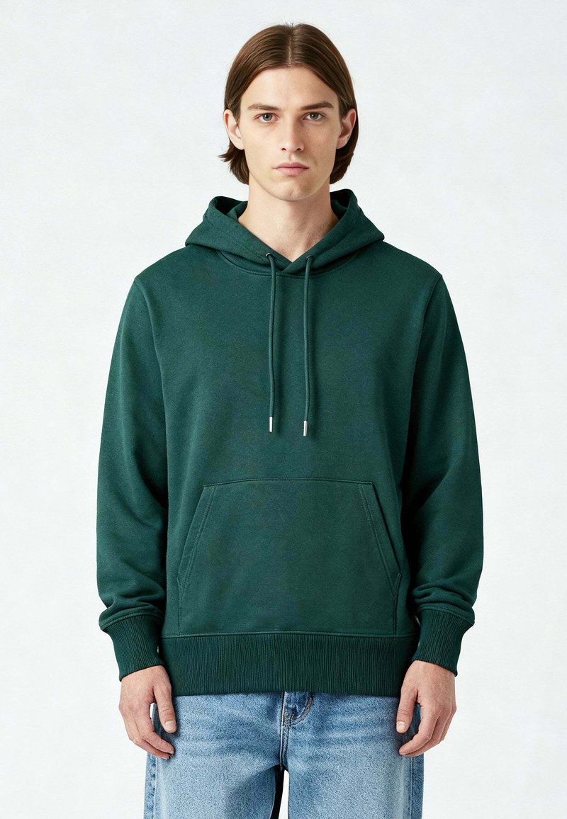 Jeune homme aux cheveux bruns mi-longs portant un sweat à capuche vert foncé et un jean bleu clair, debout devant un fond uni.