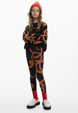 Zwarte sweatshirt met rode en gele draaiende patronen, zwarte leggings met bijpassende ontwerpen en een helderrode beanie. Dikke witte loafers.