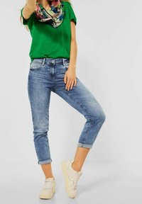 Zelené tričko s krátkým rukávem v kombinaci s světle modrými zkrácenými džíny. Džíny mají opraný vzhled a ohrnuté nohavice, doplněné béžovými teniskami.