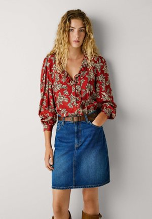 Blusa rossa floreale con scollatura arricciata, abbinata a una gonna di jeans blu aderente e una cintura marrone. L'outfit presenta un tessuto testurizzato e motivi distintivi.