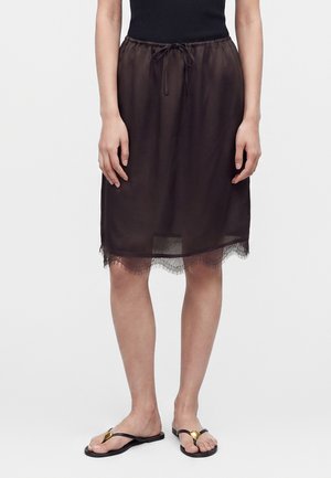 Kobieta nosząca czarną spódnicę do kolan z koronkową lamówką, dopasowaną czarną bluzkę i czarne klapki z złotymi serduszkami.