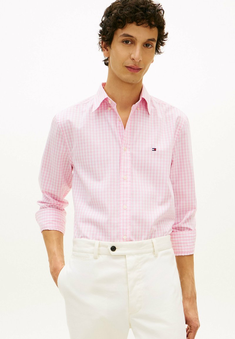 Homme portant une chemise à carreaux rose clair à manches retroussées, pantalon blanc, une main dans la poche, fond neutre.