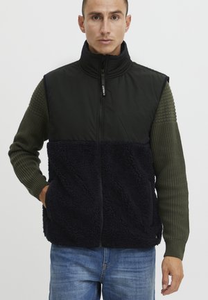 Ærmeløs vest med en overdel i sort nylon og en nederdel i navy fuzzy fleece, kombineret med ærmer i olivengrøn struktureret strik. Lynlås lukning.
