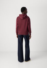 Felpa bordeaux con cappuccio regolabile, polsini e fondo a costine; abbinata a jeans dark a gamba larga e scarpe beige.