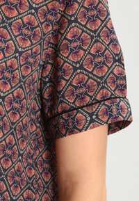 Kurzarmshirt mit geometrischem Muster in Orange, Lila und Beige auf dunkler Stoffbasis, zeigt einen Teil eines nackten Arms vor einem hellen Hintergrund.