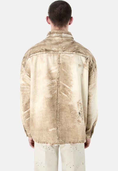 Un homme de dos porte une veste en velours côtelé beige usée et un pantalon blanc cassé avec des taches sombres mouchetées, sur un fond uni.