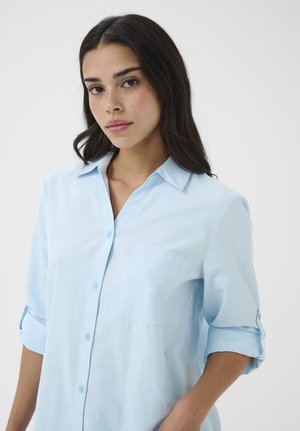 Mladá žena s dlhými tmavými vlasmi, ktorá má na sebe svetlomodrú košeľu na gombíky s vyhrnutými rukávmi, na jednoduchom bielom pozadí.