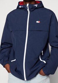 Námořnická větrovka s bílými zipy a detaily. Má nastavitelnou kapuci a elastické manžety. Logo je umístěno na hrudi.