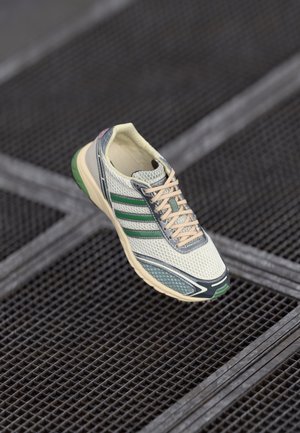 Hvid og grøn atletiksneaker med beige snørebånd, der hviler på et mørkt metalristegulv med et gittermønster.
