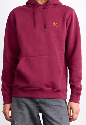 Personne portant un sweat à capuche Adidas rouge foncé avec poche avant et logo orange, associé à un jean gris.