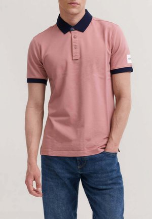 Uomo che indossa una polo rosa a maniche corte con colletto e polsini blu navy, abbinata a jeans blu, in piedi con una mano in tasca.