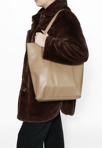 Borsa a tracolla in pelle beige con una forma strutturata, texture liscia e manici corti, tenuta da una persona che indossa un cappotto di pelliccia marrone.