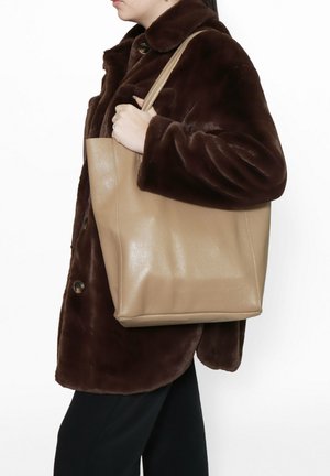 Beige leather tote bag with a structured shape, smooth texture, and short shoulder straps, held by a person wearing a brown fur coat.
