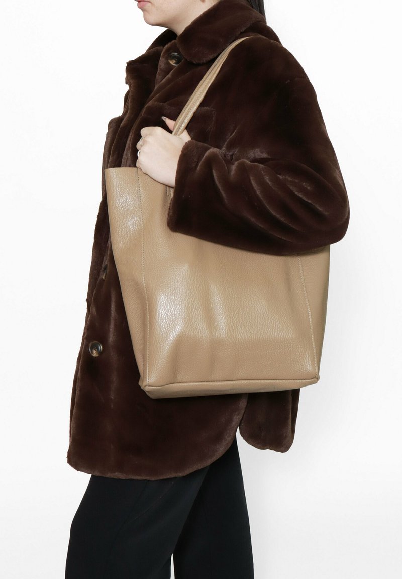 Borsa a tracolla in pelle beige con una forma strutturata, texture liscia e manici corti, tenuta da una persona che indossa un cappotto di pelliccia marrone.