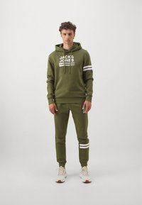 Olivgrön hoodie med vit logotyp och ränder på ärmarna, kombinerad med matchande joggers. Materialet verkar mjukt; minimalistisk design, avslappnad passform.