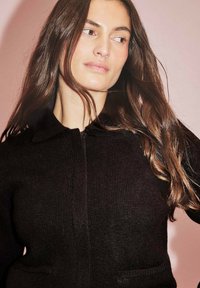 Melnā adīta jakā ar apkakli un priekšējām rāvējslēdzēja aizdari, divas sānu kabatas un ribota tekstūra uz rozā fona.