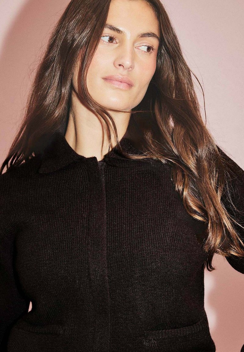 Melnā adīta jakā ar apkakli un priekšējām rāvējslēdzēja aizdari, divas sānu kabatas un ribota tekstūra uz rozā fona.