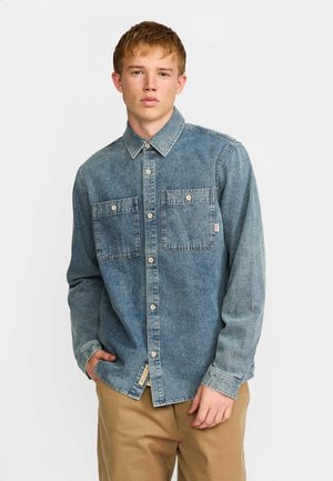 Jeune homme debout portant une chemise en jean bleue à boutons avec deux poches poitrine et un pantalon beige, sur un fond clair uni.