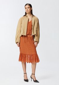 Giubbotto bomber beige con maniche a palloncino sopra un vestito senza maniche in rust drappeggiato con orlo in pizzo. Tacchi neri con fasce completano il look.