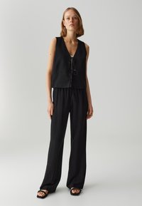 Gilet nero in lino con scollo a V e chiusura con cravatta, abbinato a pantaloni neri larghi e sandali flat, su uno sfondo neutro.