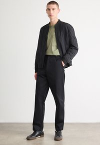 Veste bomber noire avec des poignets côtelés, t-shirt vert avec poche et pantalon noir. Les baskets ont des lacets foncés et une semelle texturée.