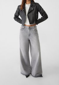 Giacca cropped in pelle nera con zip, abbinata a jeans grigi a gamba larga con vita alta e lievi dettagli di scoloritura.