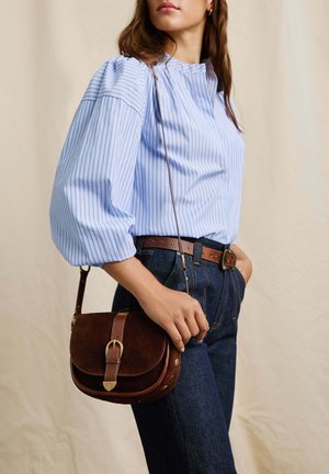 Brown suede crossbody bag with a gold buckle, paired with a light blue striped blouse and dark denim high-waisted jeans.