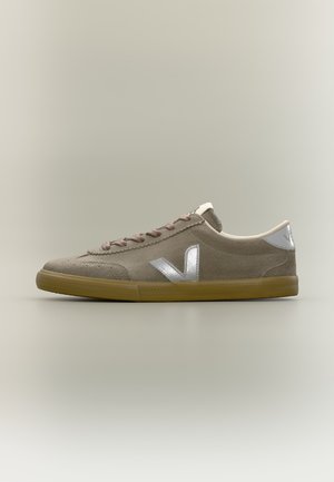 Zapatilla beige de ante de corte bajo con cordones marrones, logo metálico plateado "V" en el lateral, lengüeta reflectante en el talón y suela de goma.