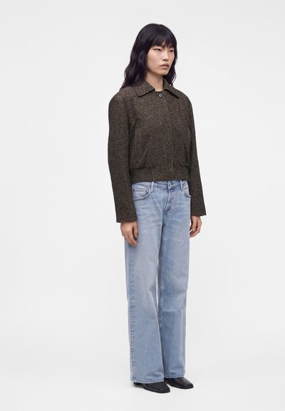 Femme portant une veste texturée marron foncé, un jean large bleu clair et des chaussures noires pointues, debout devant un fond blanc uni.