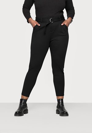 Persona que lleva pantalones negros de tiro alto con cinturón y botines negros, con las manos en los bolsillos, de pie frente a un fondo sencillo.