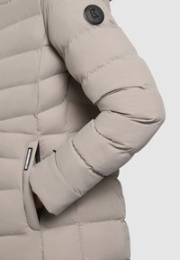 Doudoune beige clair avec un design matelassé, arborant un patch logo noir sur la manche et un détail de poche zippée. Texture lisse.
