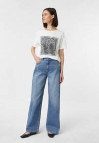 Weißes T-Shirt mit einem schwarz-weißen grafischen Quadratdruck, kombiniert mit hellblauen weiter geschnittenen Jeans und schwarzen Ballerinas.