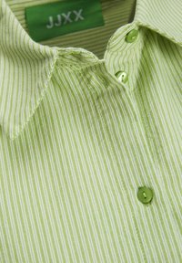 Camicia a righe verde chiaro con colletto a punta, dotata di bottoni verdi. Il tessuto ha una texture sottile e un'etichetta che riporta "JJXX."