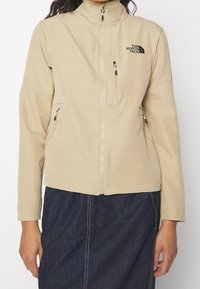 Beige zip-up jacka tillverkad av mjukt tyg, med hög krage, två sidofickor och en bröstficka med en svart logotyp.