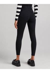 Černé skinny džíny s vysokým pasem, hladkou texturou, zadními kapsami a přiléhavým střihem. V kombinaci s černými botami na světlém pozadí.