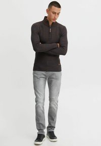 Dunkel gestreifter Pullover mit kurzem Reißverschlusskragen, gerippter Textur und einem kleinen braunen Etikett. Getragen mit hellgrauen Jeans und schwarzen Sneakern.