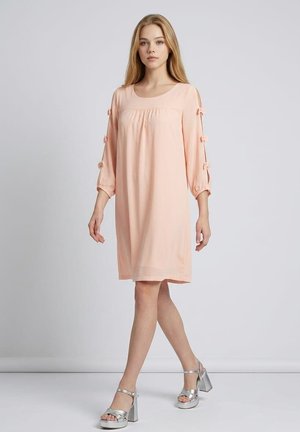 Mujer joven con un vestido rosa claro hasta la rodilla con mangas recortadas atadas con lazos y sandalias plateadas de tacón alto.