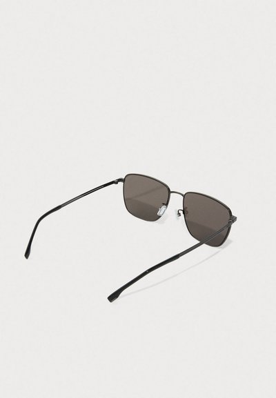 Lunettes de soleil à monture noire en métal avec des branches fines et des verres rectangulaires sombres reposant sur une surface blanche, vues de dessus.