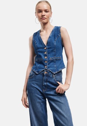 Farkkutakki, jossa on V-kaulussuunnittelu, kuusi nappia edessä, kaksi sivutaskua ja kontrastiväriset tikkaukset. Puettu farkkuhousujen päälle.