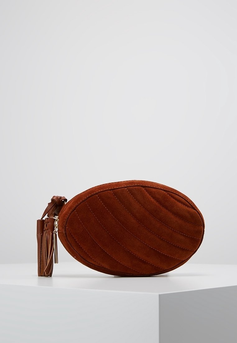 Pochette ovale en daim matelassé marron avec fermeture éclair et porte-clés en cuir à pompon, posée sur un piédestal blanc sur fond clair uni.