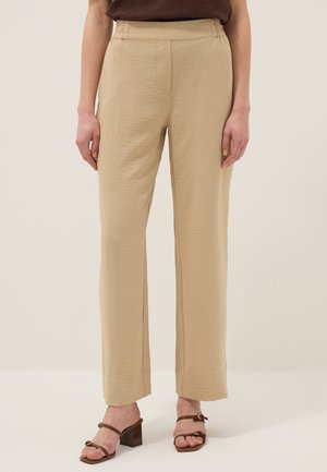 Beige, gestructureerde, rechte broek gecombineerd met bruine sandaal met bandjes en blokhakken, gedragen door een staand persoon tegen een effen achtergrond.