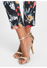 Sandales à talons dorés avec un design à bout ouvert, featuring une fine bride de cheville et un talon aiguille texturé. Pantalons à motif floral visibles au-dessus.