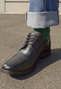Zwart leren schoenen met een gladde afwerking, vetersluiting en een lichtpuntige neus. Draagt zich met blauwe denim, opgerolde broek en groene sokken.